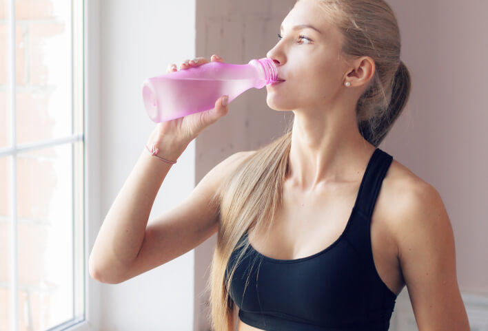 Woman in athletic wear drinking from a pink bottle indoors.